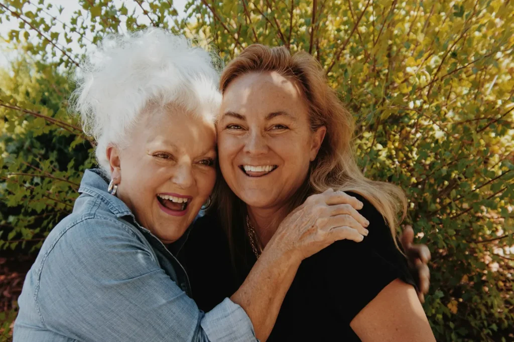 An older woman with white hair and her adult daughter hug and laugh together in a sunny garden