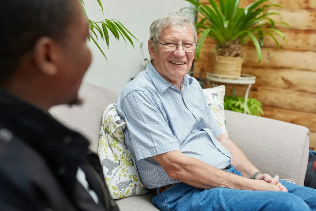 An older man in a striped shirt laughs while chatting with a support worker on a couch at home