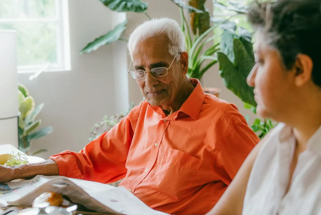An older man sits at a sunlit dining table going through papers with a younger woman beside him