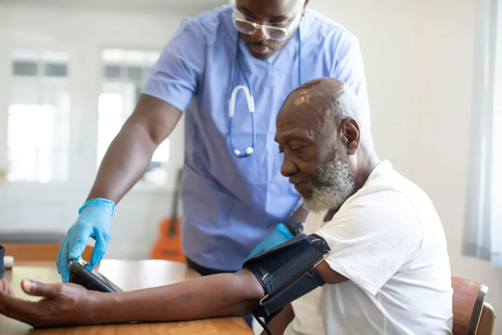A nurse in blue scrubs checks the blood pressure of an older man seated at a table at home