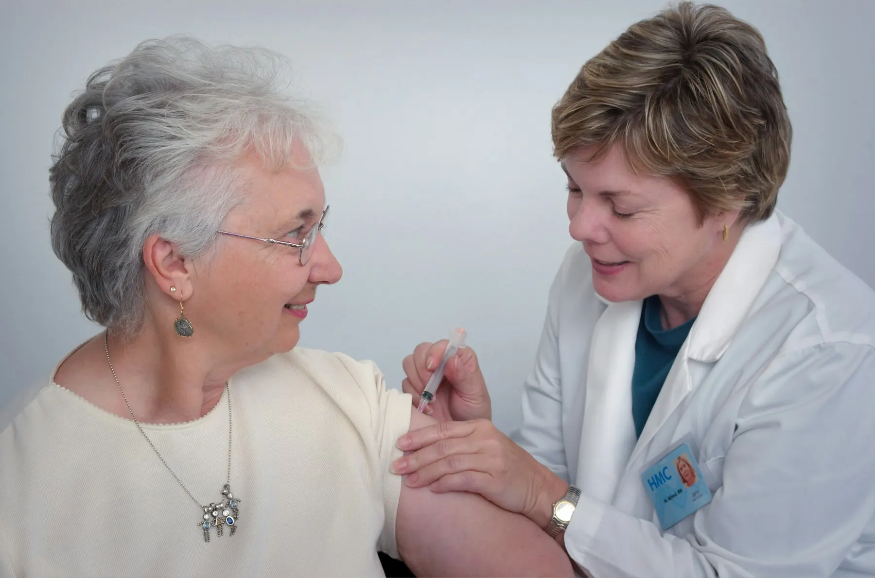 A nurse in a white coat gives a smiling older woman with grey hair an injection in her upper arm