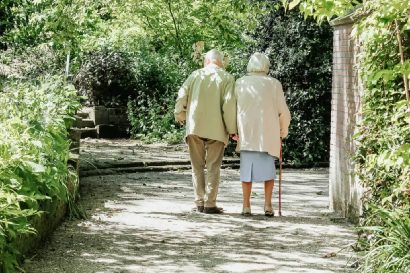 An older couple walk arm in arm along a leafy garden path, the woman using a walking stick
