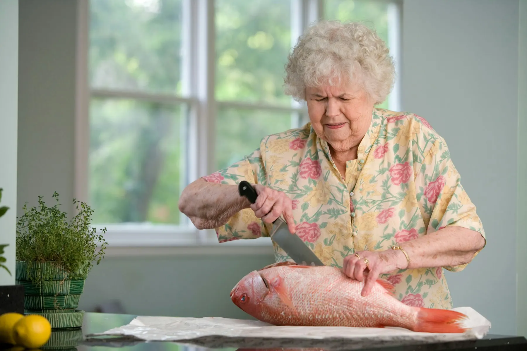 An older woman with curly white hair cuts a whole red fish with a large knife at her kitchen bench