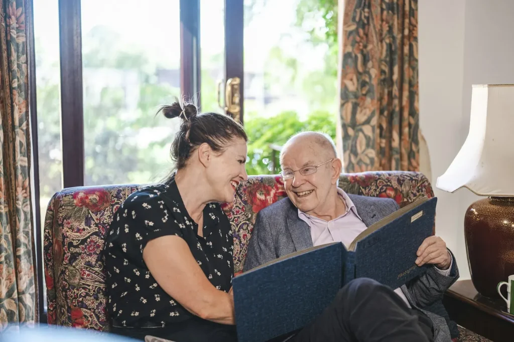 An older man and a woman laugh together as they look through a folder on a couch at home