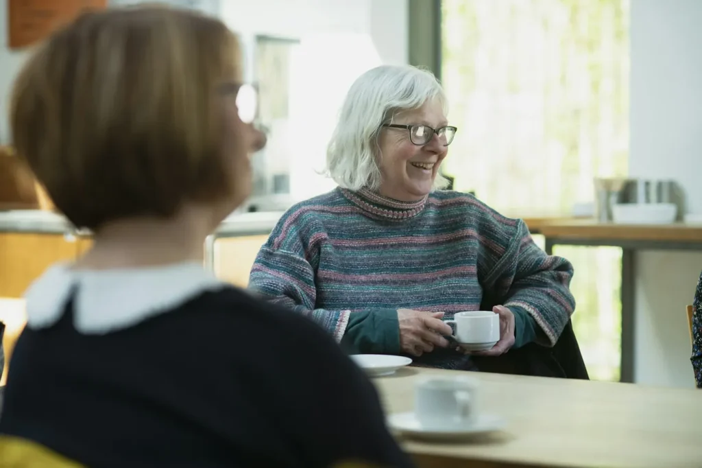 An older woman with a cup of tea smiles while chatting with a companion at a table by the window