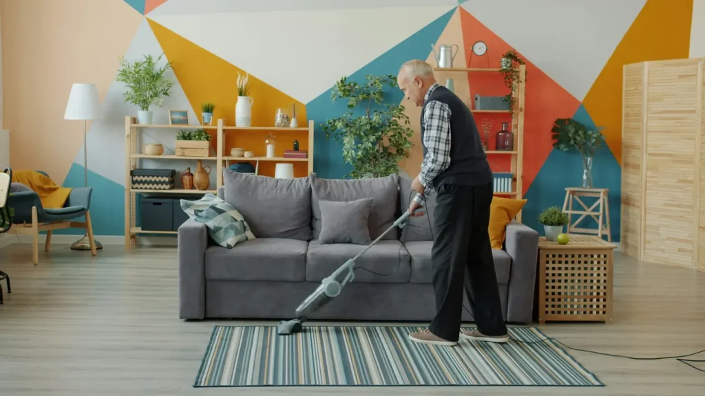 An older man vacuums the rug in front of the couch in a bright living room