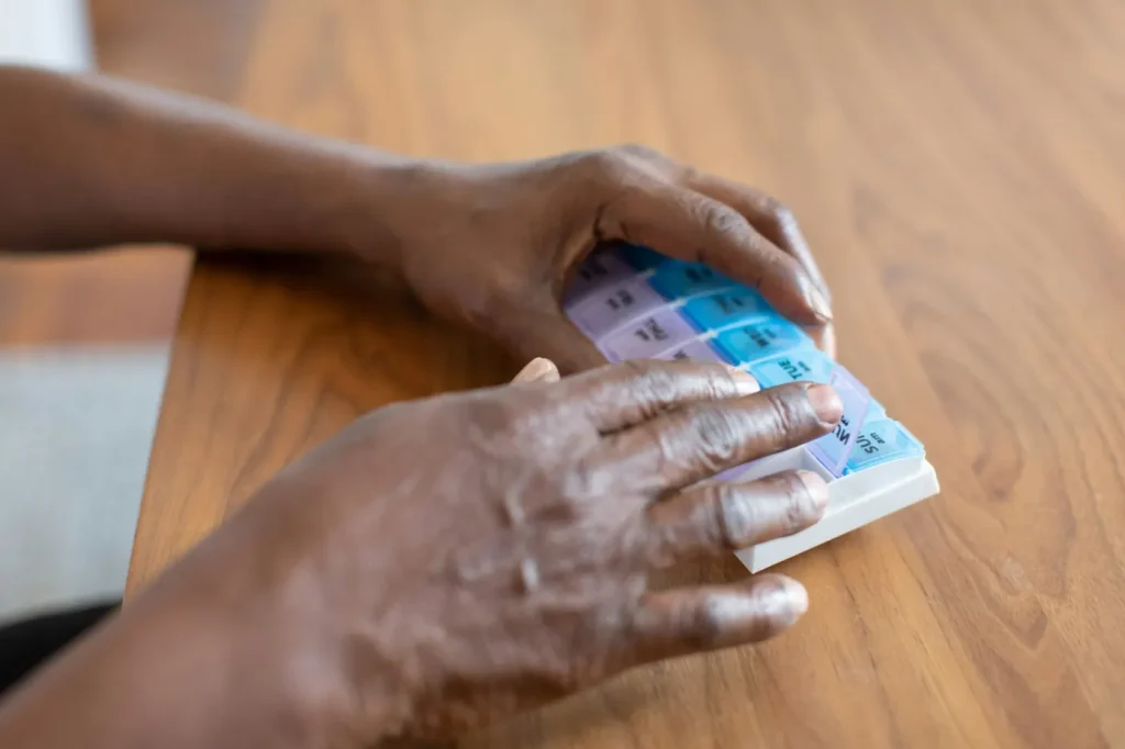 An older person’s hands open a compartment of a weekly pill organiser on a timber table