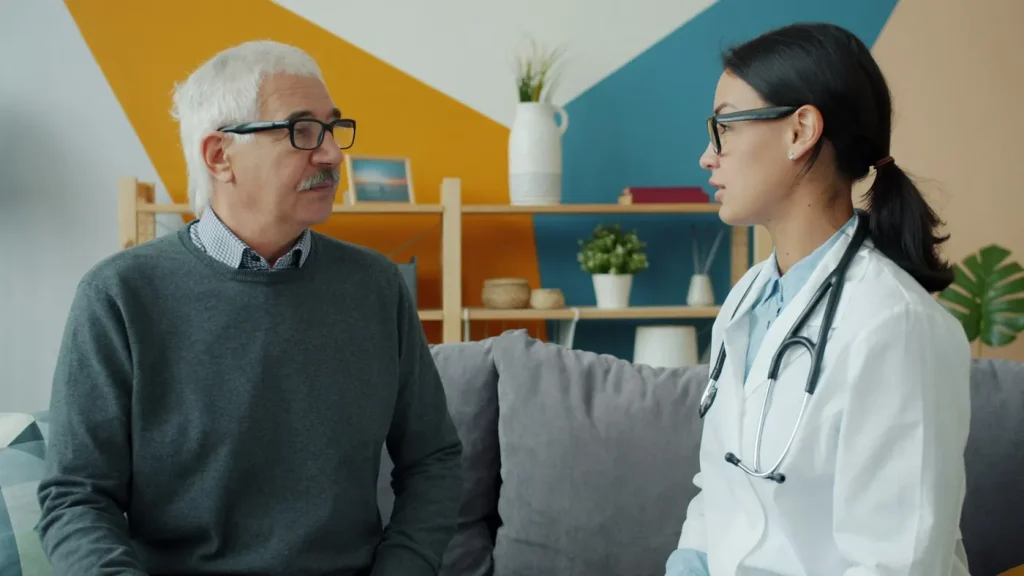 A clinician with a stethoscope talks with an older man on the couch in his living room