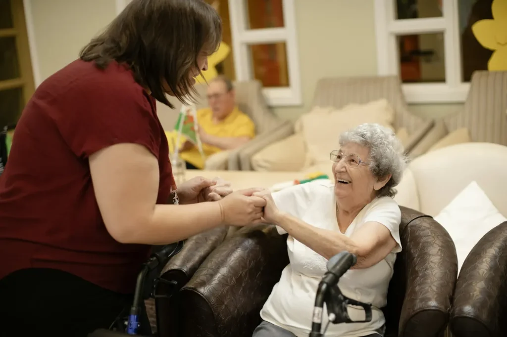 A care worker holds both hands of a smiling older woman to help her rise from her armchair