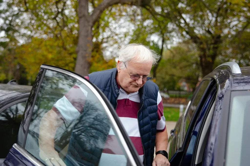 An older man opens the door to step into a car on a tree-lined street